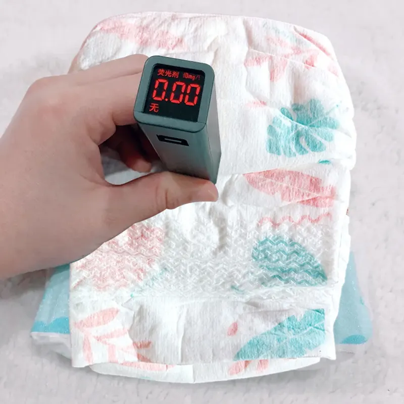 Hand testing a baby diaper with a fluorescence detector; display reads 0.00, indicating no migratory optical brighteners.